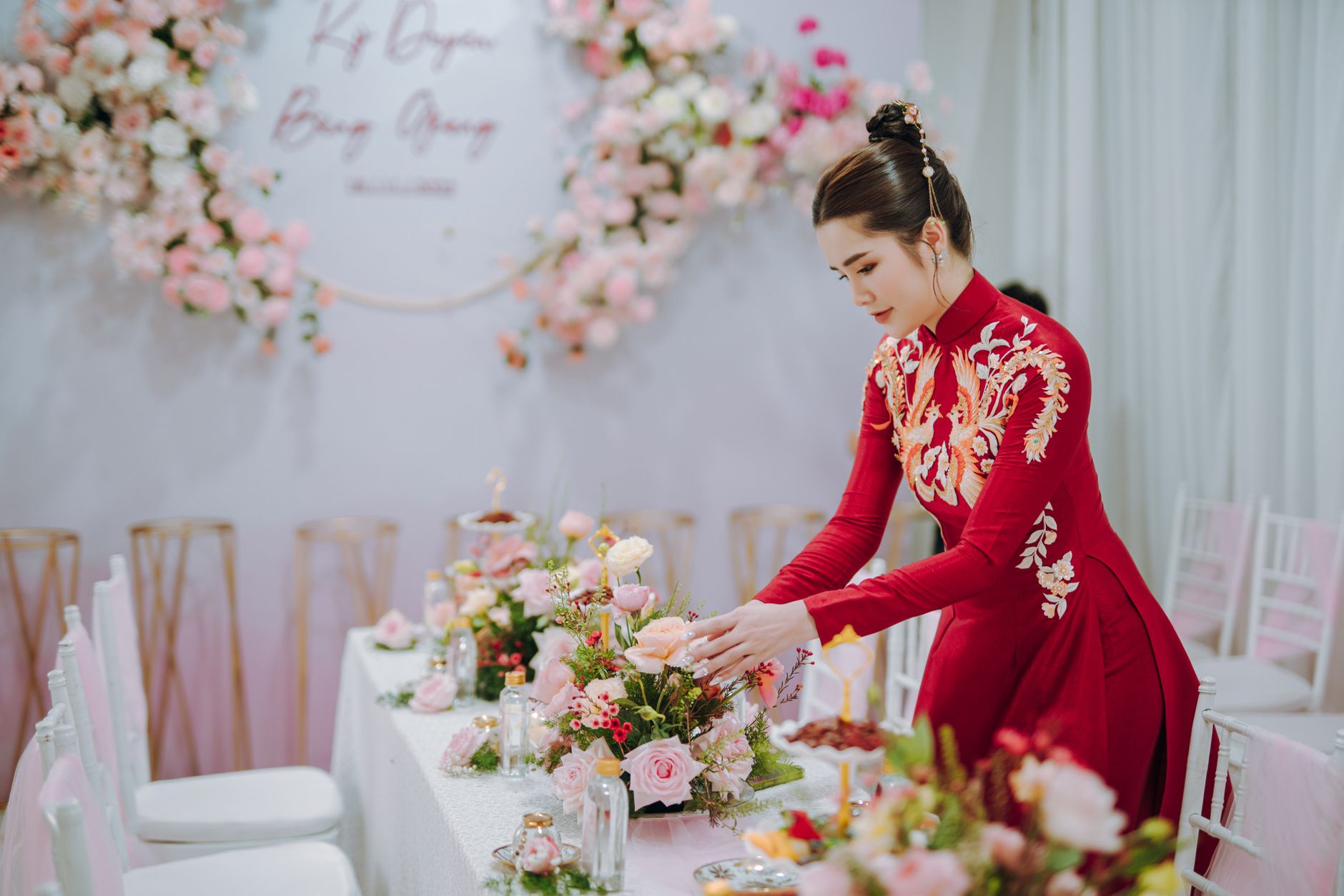 An Asian woman in traditional red dress arranges a floral centerpiece at an elegant indoor event.