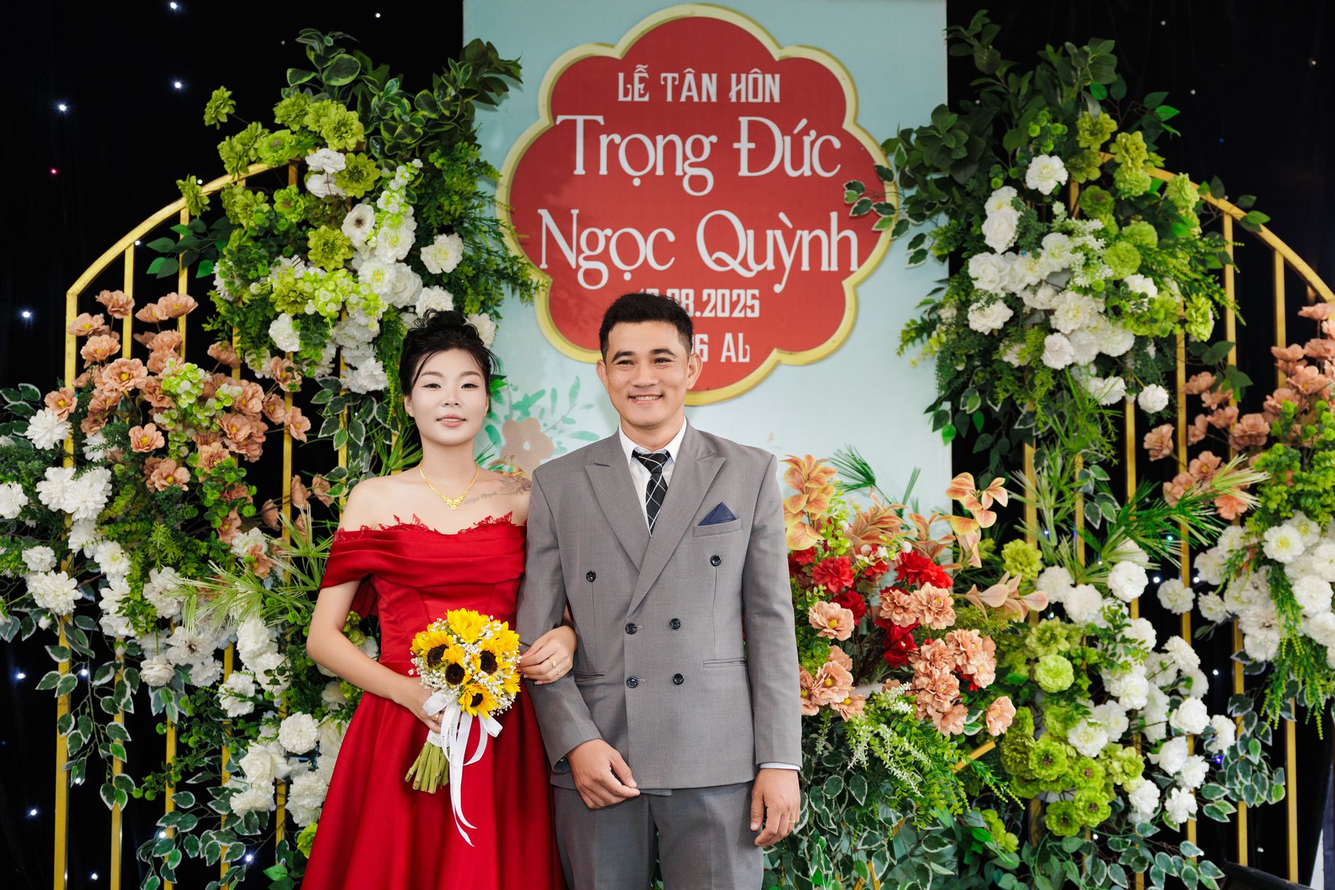 A couple poses at a wedding with a vibrant floral backdrop and a celebratory sign.