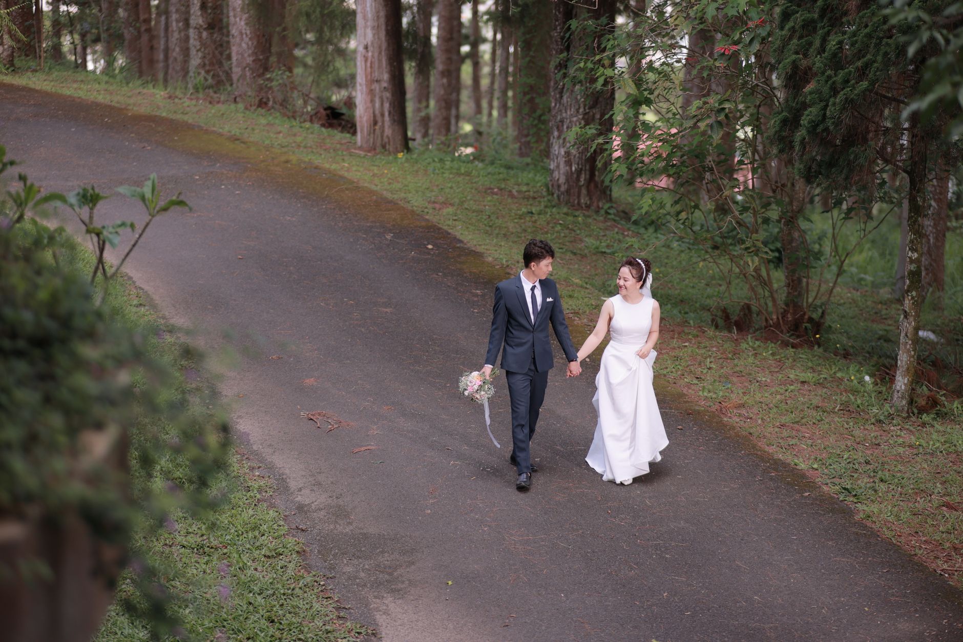 Bride and groom holding hands, walking through a forest pathway in Vietnam.